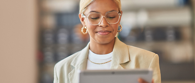 Женщина в очках и с серьгами, в легкой куртке сосредоточенно читает контент на планшете.