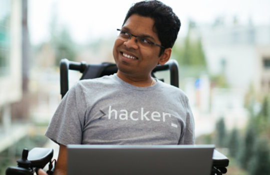 A man who uses a wheelchair wears a hacker t-shirt and looks to the side.