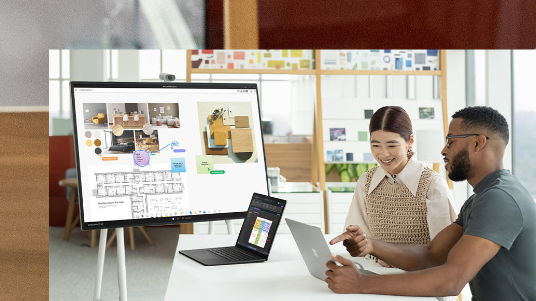 Dua orang rakan sekerja berinteraksi dengan dua peranti Surface Laptop 5 dan Hub 2S di latar belakang
