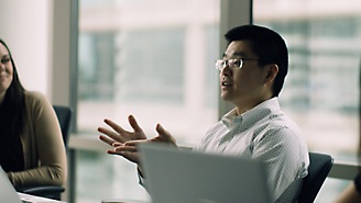 Um homem usando óculos gesticulando enquanto fala ao lado de seu laptop em um ambiente de escritório