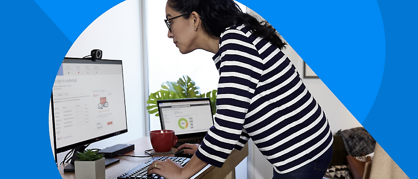 줄무늬 셔츠를 입은 한 여성이 컴퓨터 모니터 두 대가 있는 책상에서 키보드를 치며 일하고 있습니다.