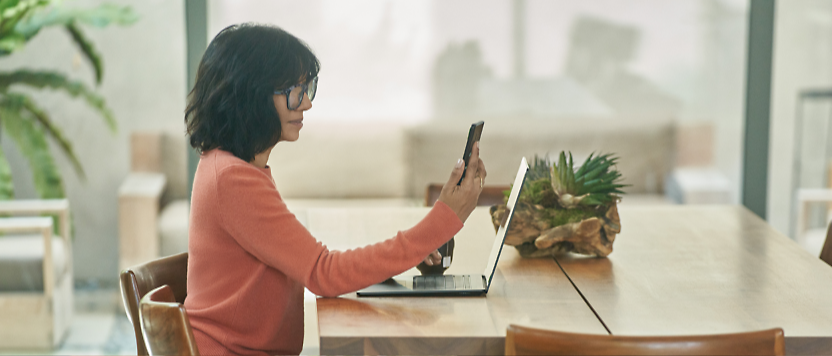 אישה ממושקפת משתמשת במחשב נישא ובטלפון חכם ליד שולחן עץ מודרני ועליו צמח בעציץ 