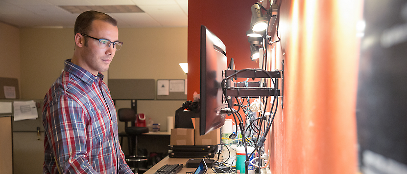 Ekose gömlek giyen gözlüklü bir adam, dağınık bir ofis ortamında bilgisayar ekranına odaklanıyor.