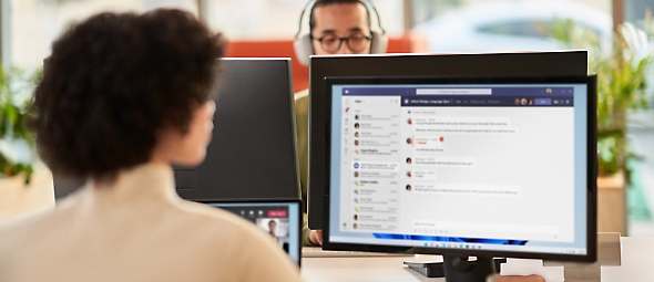 אישה משתפת פעולה במהלך פגישת Microsoft Teams בזמן עבודה על רקע משרד פתוח בצג כפול