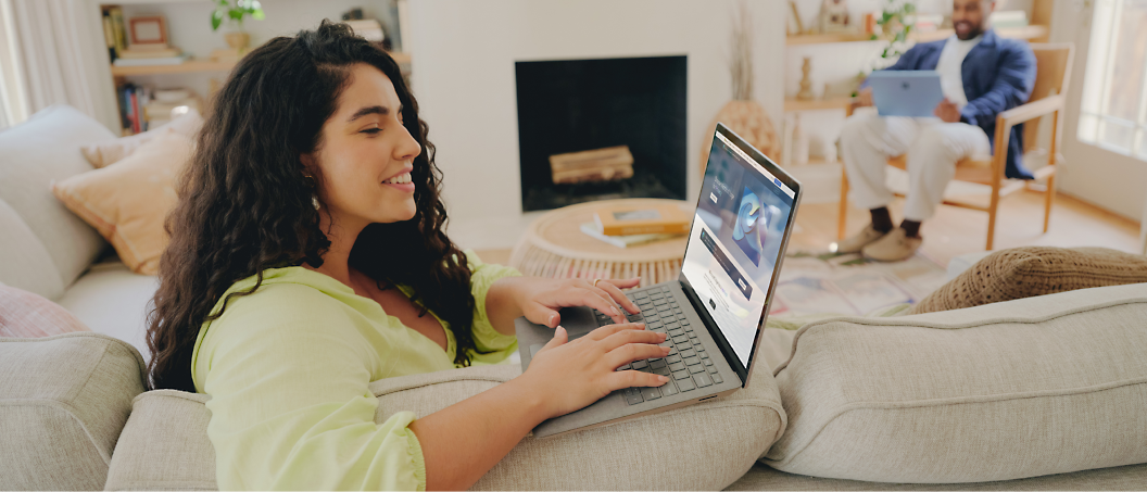 A young woman smiling and using a laptop on a couch, video chatting with friends, with another person 