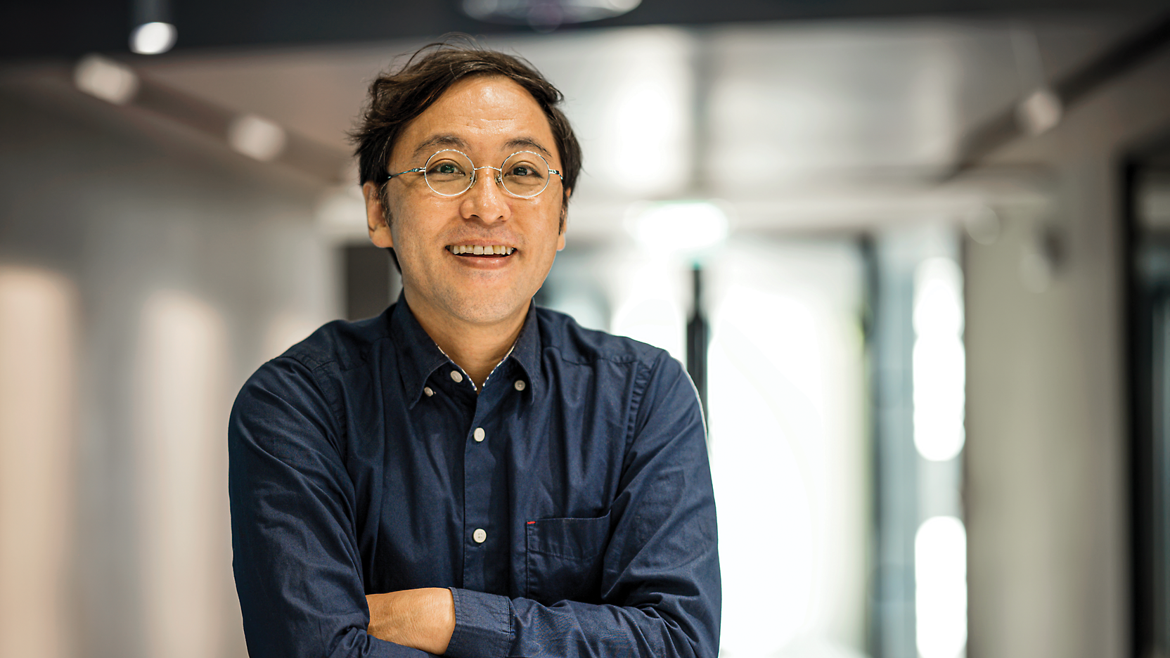 A middle-aged asian man in a blue shirt smiling confidently, arms crossed, standing in an office corridor.