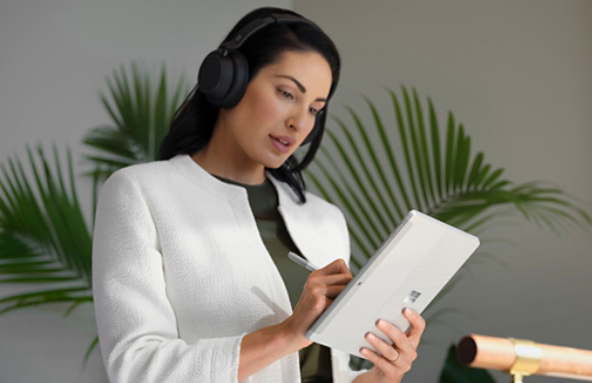 คนสวม Surface Headphones 2+ คนหนึ่งกำลังใช้ปากกา Surface นำทาง Surface Go 4 ในโหมดแท็บเล็ตอยู่ที่ทำงาน