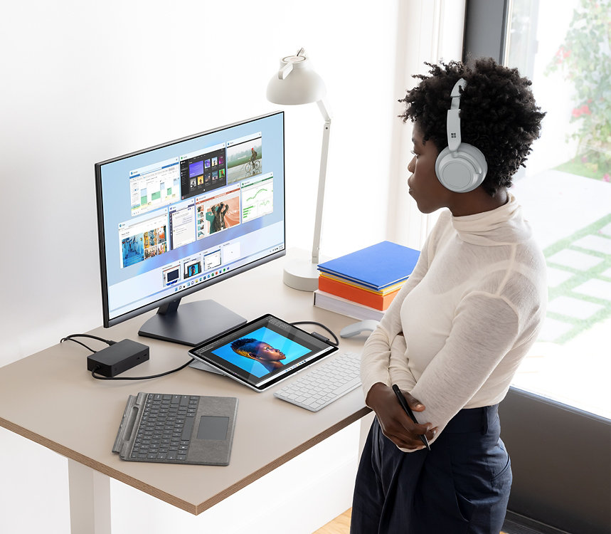 Eine Person mit Surface Headphones steht an einem Tisch mit einem Surface-Gerät, einer Type Cover-Tastatur und einem Monitor.