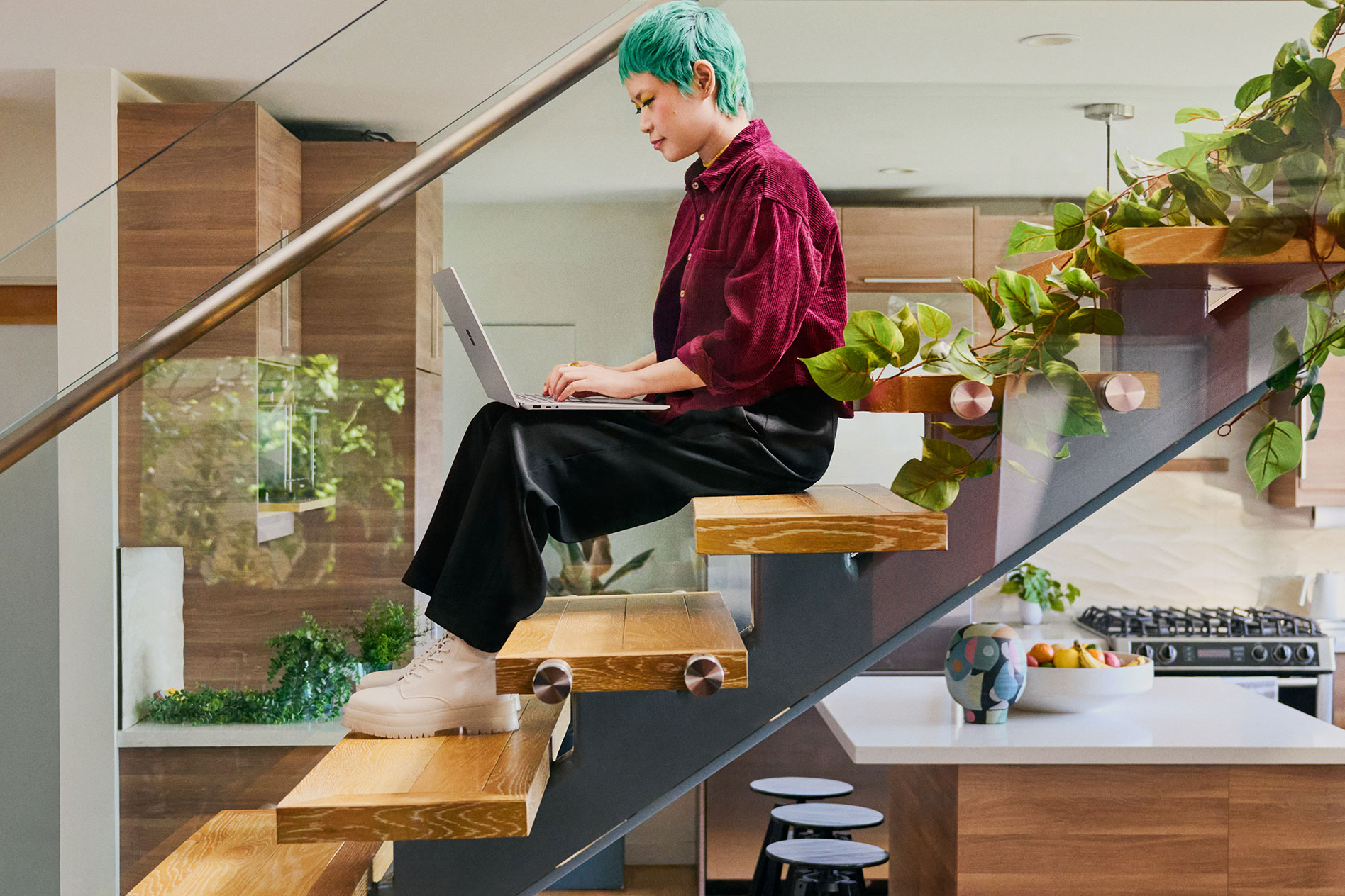 Seorang pemuda yang duduk di atas tangga di dapur moden menaip pada Surface Laptop 5 miliknya.