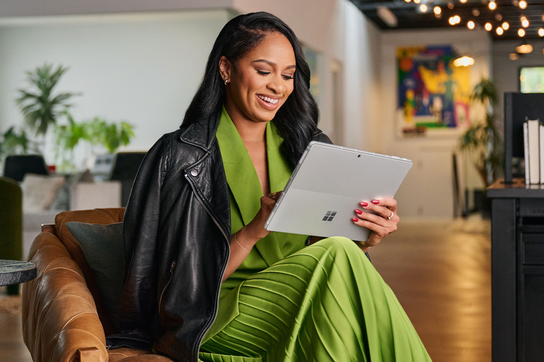 Seorang wanita yang memakai gaun hijau panjang dan jaket kulit hitam duduk di atas kerusi sambil berinteraksi dengan Surface Pro 9 miliknya dalam mod tablet.
