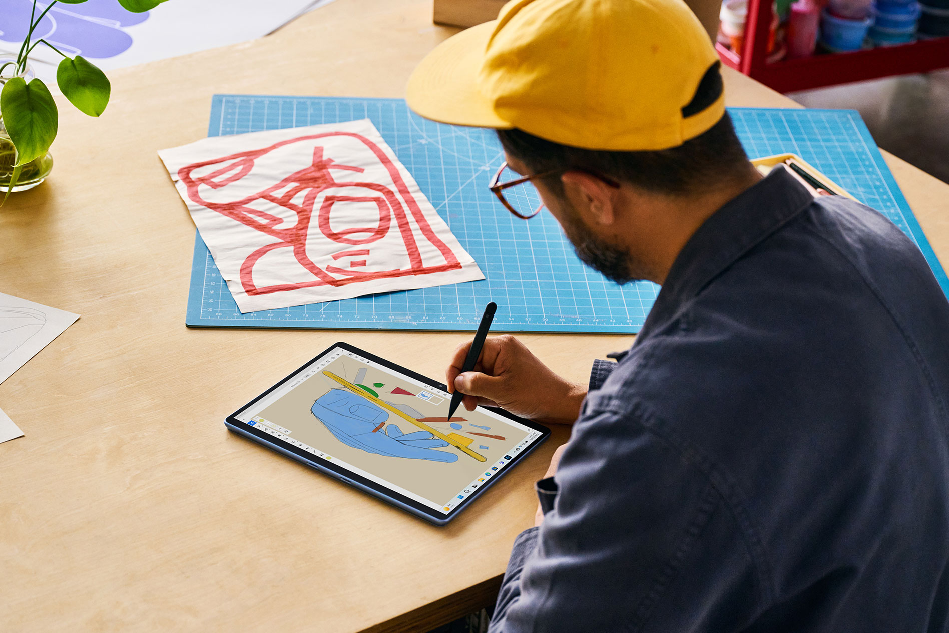 Seorang lelaki yang memakai topi besbol duduk di tepi meja kayu dan melukis dengan Surface Slim Pen 2 pada Surface Pro 9 miliknya dalam mod tablet.