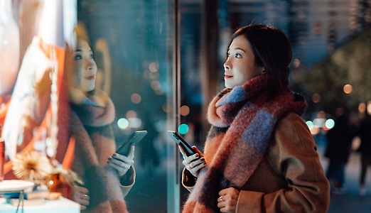 Een persoon met een grote sjaal die een mobiele telefoon vasthoudt en in een winkeletalage kijkt die is verlicht door de stadslampen achter de persoon.