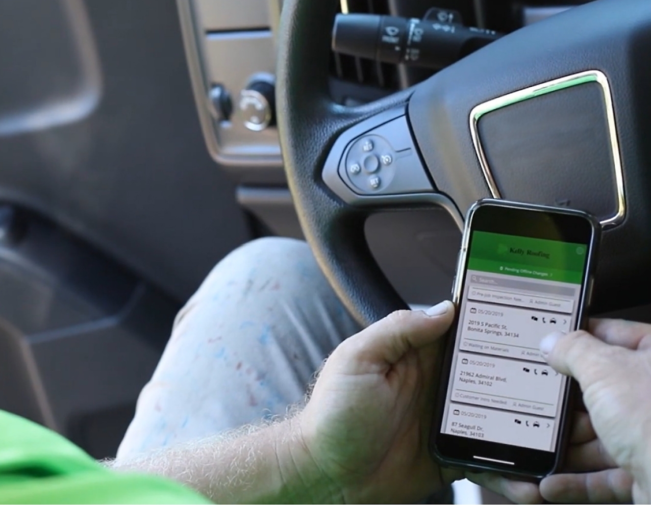 Egy zöld pólóban lévő férfi mobiltelefont használ egy teherautó vezetése közben.