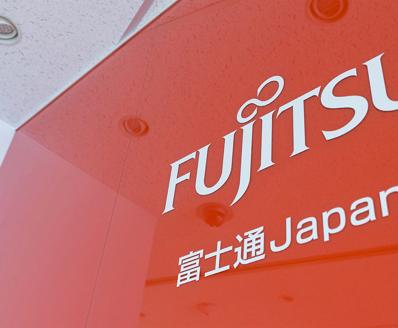 Primer plano parcial de un cartel de Fujitsu con logotipo y texto en japonés sobre un fondo rojo, perspectiva en ángulo 