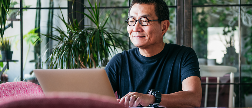 Middle-aged asian man smiling while working on a laptop at a cafe table near large windows with greenery outside.
