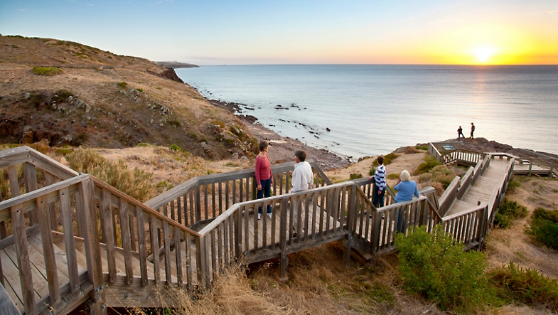 Personen die in Zuid-Australië bij zonsondergang over een houten pad in de heuvels langs de zee wandelen.