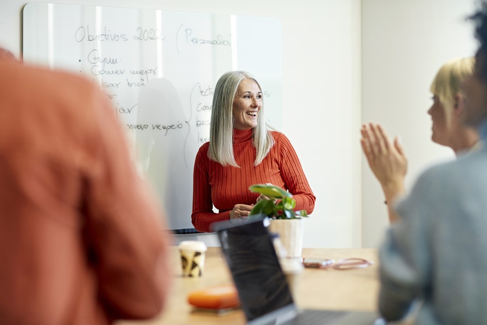 white woman with white hair and orange shirt smiling standing in front of whiteboard during team meeting