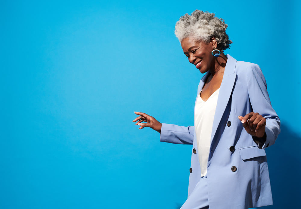 Black woman with white hair in blue suit happily step dancing against blue background