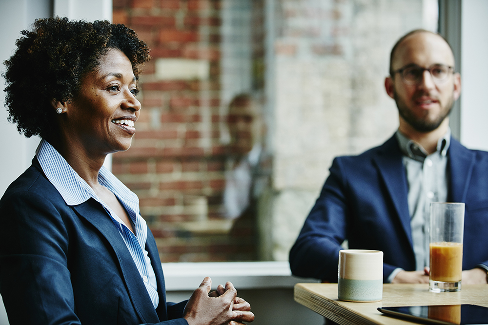 black woman smiling talking to coworkers at team lunch seated next to male colleague 