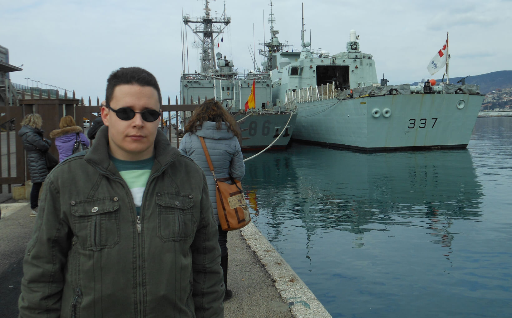 Milos standing at a harbor front with two large ships behind him.