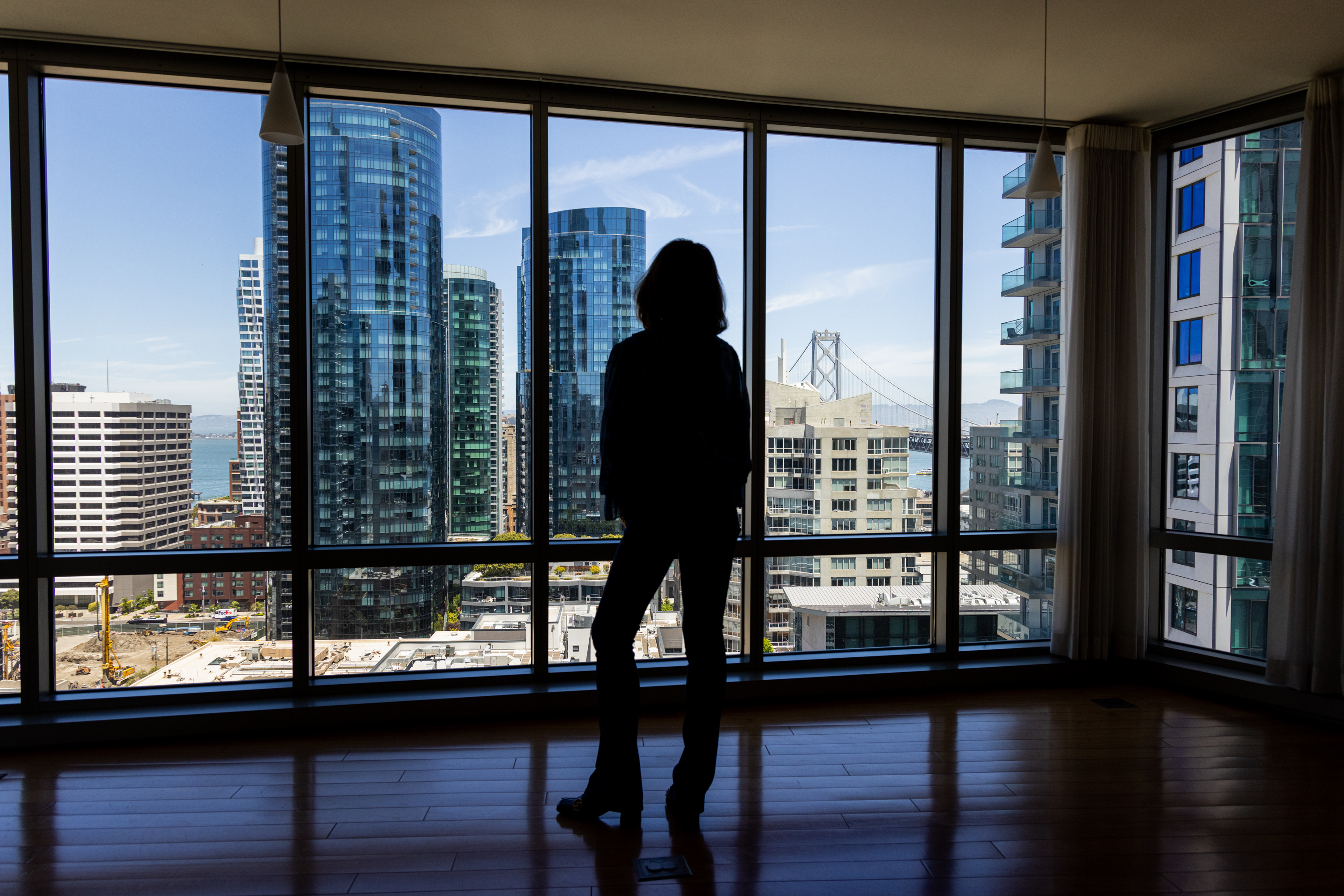 A person stands in front of a large window in an apartment, overlooking a cityscape filled with modern high-rise buildings and a partial view of a bridge.