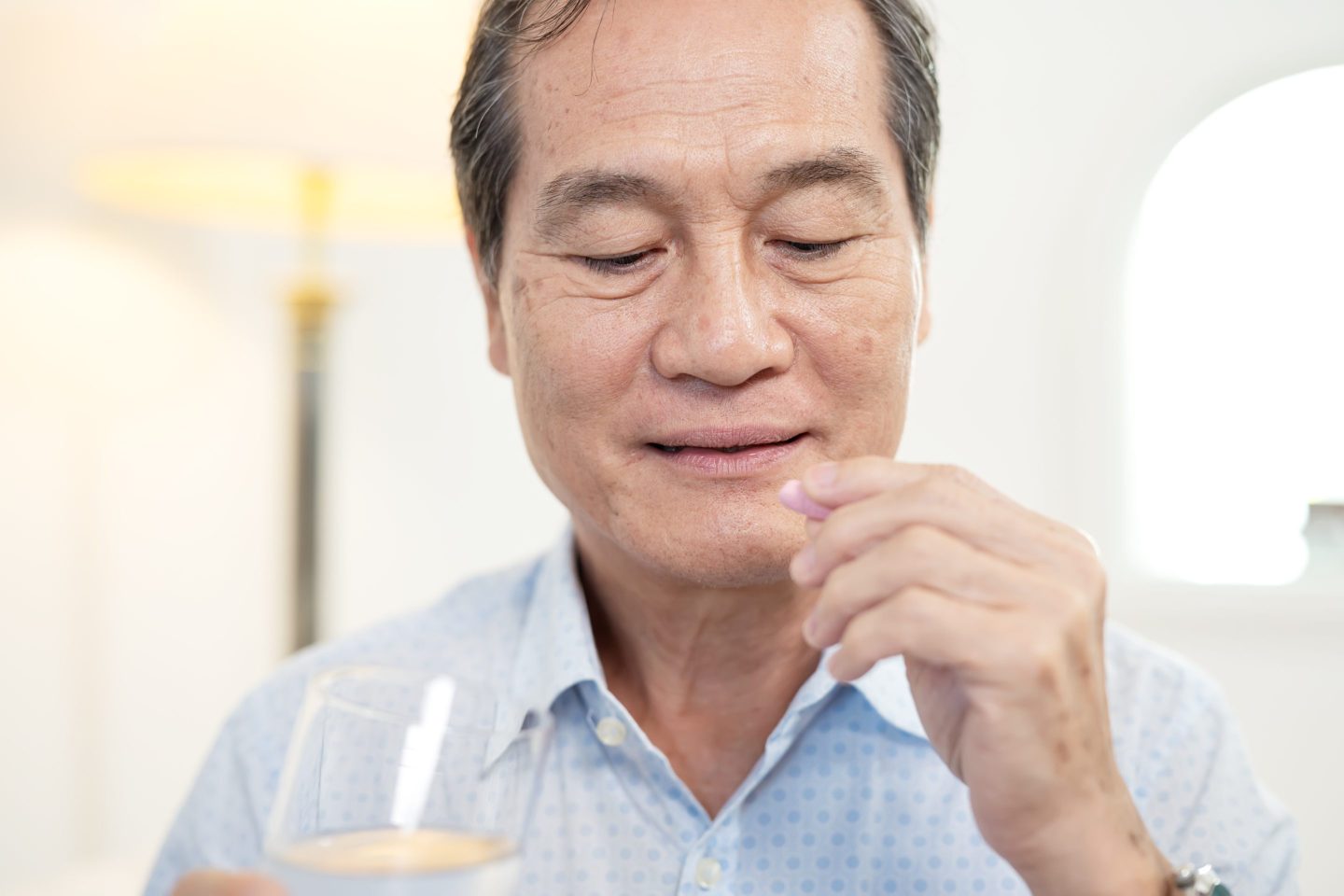 Safely Managing Medications is Essential for Senior Health. Profile view of a senior Asian male holding a glass of water and a pill in the living room at his home.