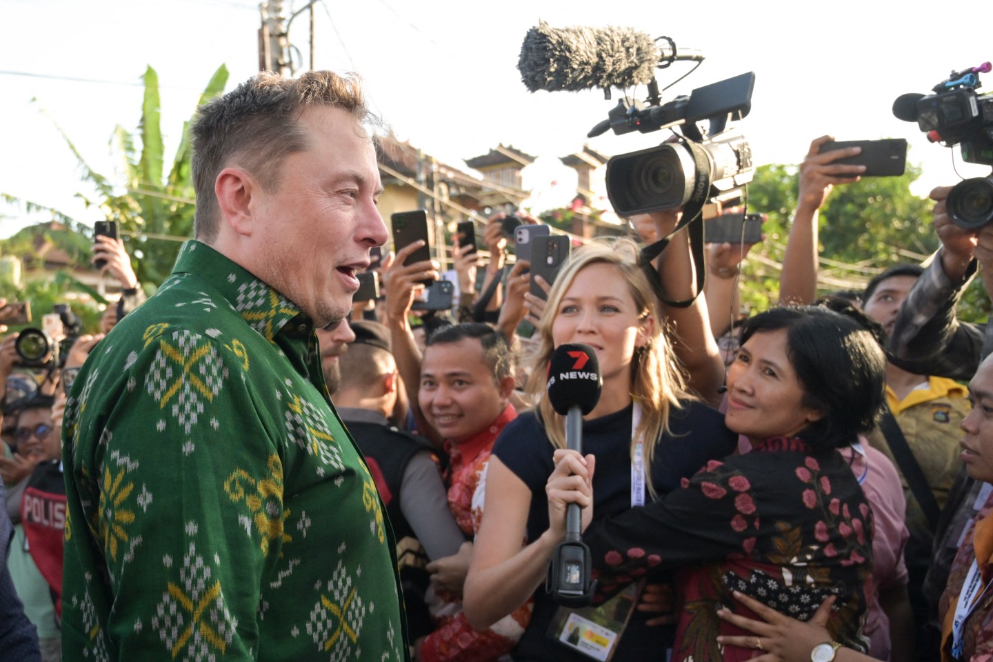 Elon Musk in profile surrounded by reporters while visiting Indonesia.