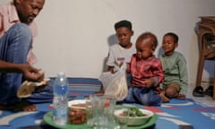 Three small children sit on the floor, while a man hands them food from a small plate.