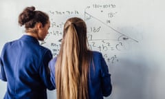 Two teenage school girls standing in front of a large whiteboard side by side solving a mathematics equation on the board