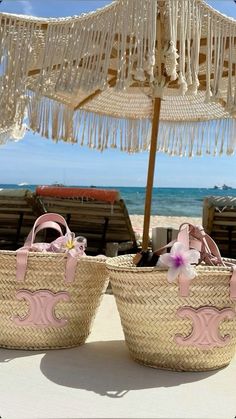 two wicker baskets with pink handles are sitting under an umbrella on the sand at the beach