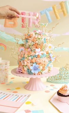 a birthday cake is decorated with pastel flowers and icing, while someone holds the letter h above it