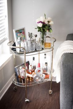 a bar cart with liquor bottles and flowers on it in front of a window next to a couch