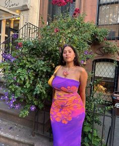 a woman is standing in front of a bush wearing a purple and orange strapless dress