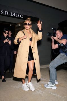 a woman in a tan trench coat and white sneakers is waving at the camera as she stands