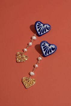 two heart shaped earrings with pearls hanging from them on a pink surface, one is gold and the other is blue