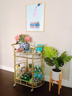 a gold bar cart with drinks on it in front of a painting and potted plant