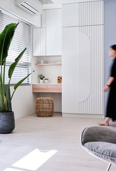 a woman is walking past a plant in a room with white walls and flooring