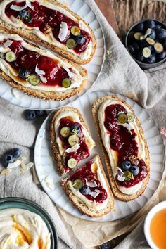 two white plates topped with sandwiches covered in jelly and blueberries next to cups of tea