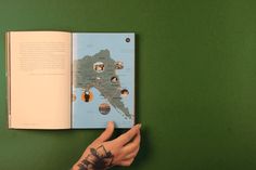 a person's hand holding an open book on top of a green surface with images of people