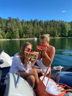 two girls eating watermelon off the back of a boat on a lake with trees in the background