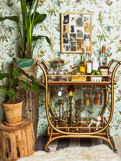 a gold bar cart with drinks on it next to a potted plant and wallpaper