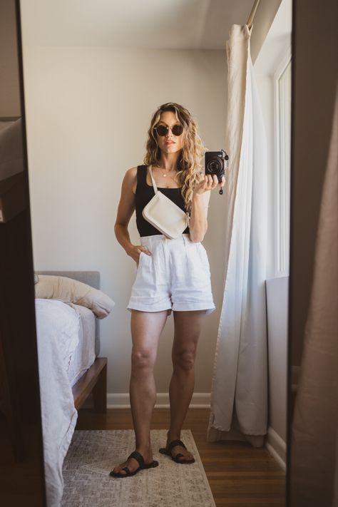 Ashley wearing a black tank, white linen shorts, black sandals, a beige belt bag, and tortoiseshell sunglasses