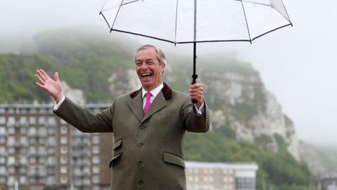 Nigel Farage pictured grinning and holding a see-through umbrella with the White Cliffs of Dover in the background