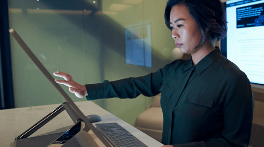 Moters, dėvinčios tamsius marškinius pritemdytame biure naršo arba dirba su „Microsoft Surface Studio“, šoninis profilis.