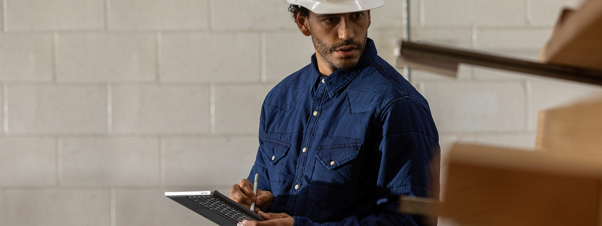 A worker is seen wearing a hardhat in an industrial setting holding a Surface Go 2 in tablet mode