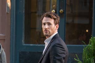 Chris Evans stands on a street in a suit, filming a scene, with a crew member in a hat and headset crouching nearby