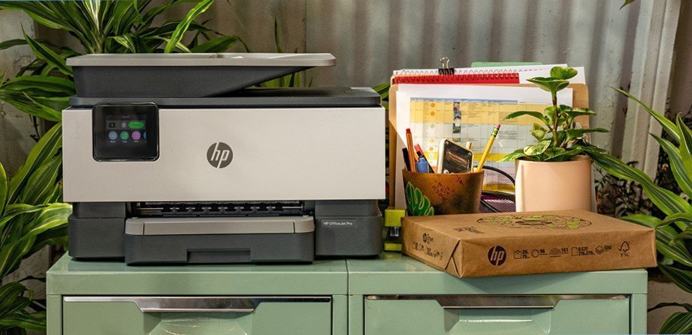 HP printer sitting on green filing cabinets next to an unopened package of paper, a cup with pens, a plant, and some documents