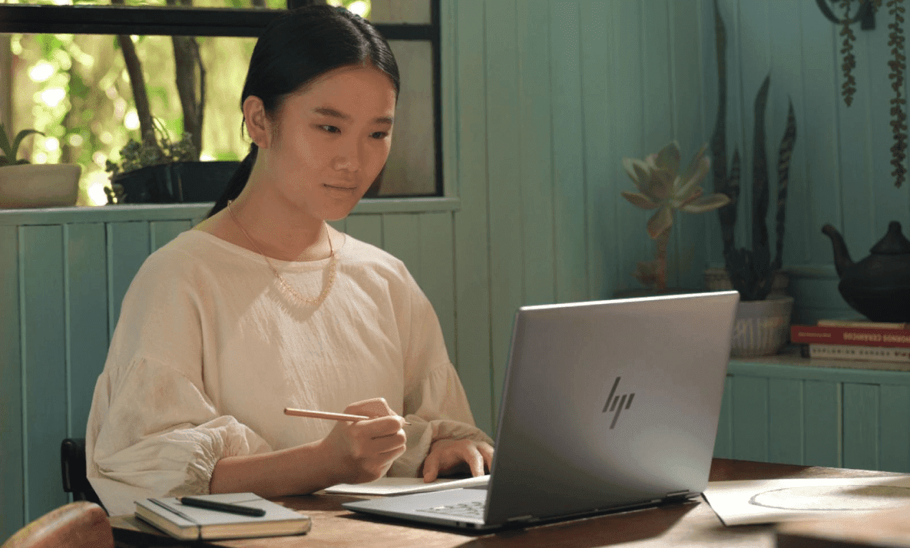Woman at a desk in a green home office, looking into an HP laptop while holding a pencil above a notebook to rec