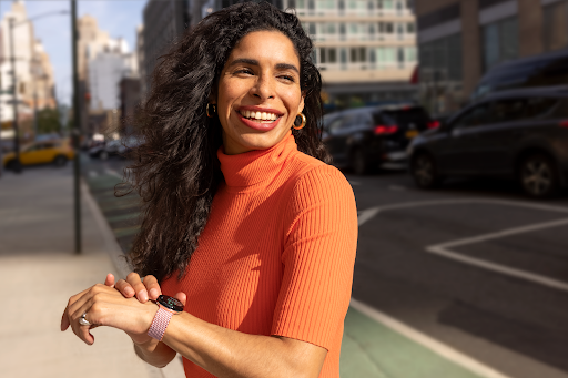 L'utilisatrice de la montre connectée se tient debout sur le trottoir d'un quartier en centre-ville. Elle sourit tout en regardant la direction indiquée sur l'application Google Maps de sa montre.