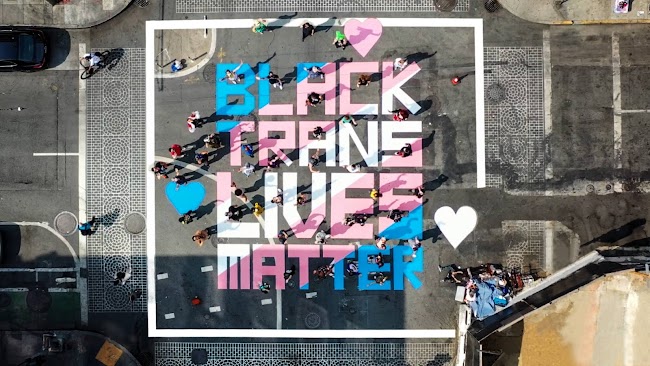 Mural de Black Trans Lives Matters pintado con spray en azul claro, rosa y blanco en intersección de calles.